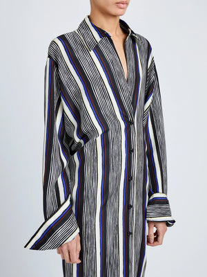 A person wears the Amira Dress by Proenza Schouler, a long-sleeved, button-down shirtdress with vertical black, white, and blue stripes in luxe Printed Silk Habotai, standing against a plain background.