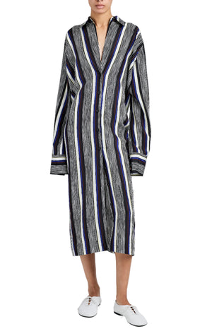 A person wearing the Amira Dress by Proenza Schouler, a loose-fitting, long-sleeve shirtdress with vertical black, white, and blue stripes in Printed Silk Habotai, paired with white lace-up shoes.