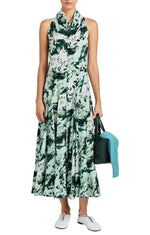 A woman wears the Proenza Schouler Adelide Dress—a sleeveless green and white patterned midi with a draped neckline and fit-and-flare silhouette—paired with white sneakers, a black bag, and a teal sweater.
