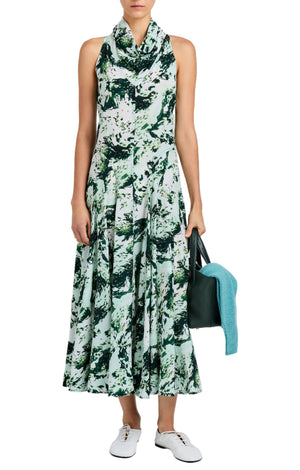 A woman wears the Proenza Schouler Adelide Dress—a sleeveless green and white patterned midi with a draped neckline and fit-and-flare silhouette—paired with white sneakers, a black bag, and a teal sweater.