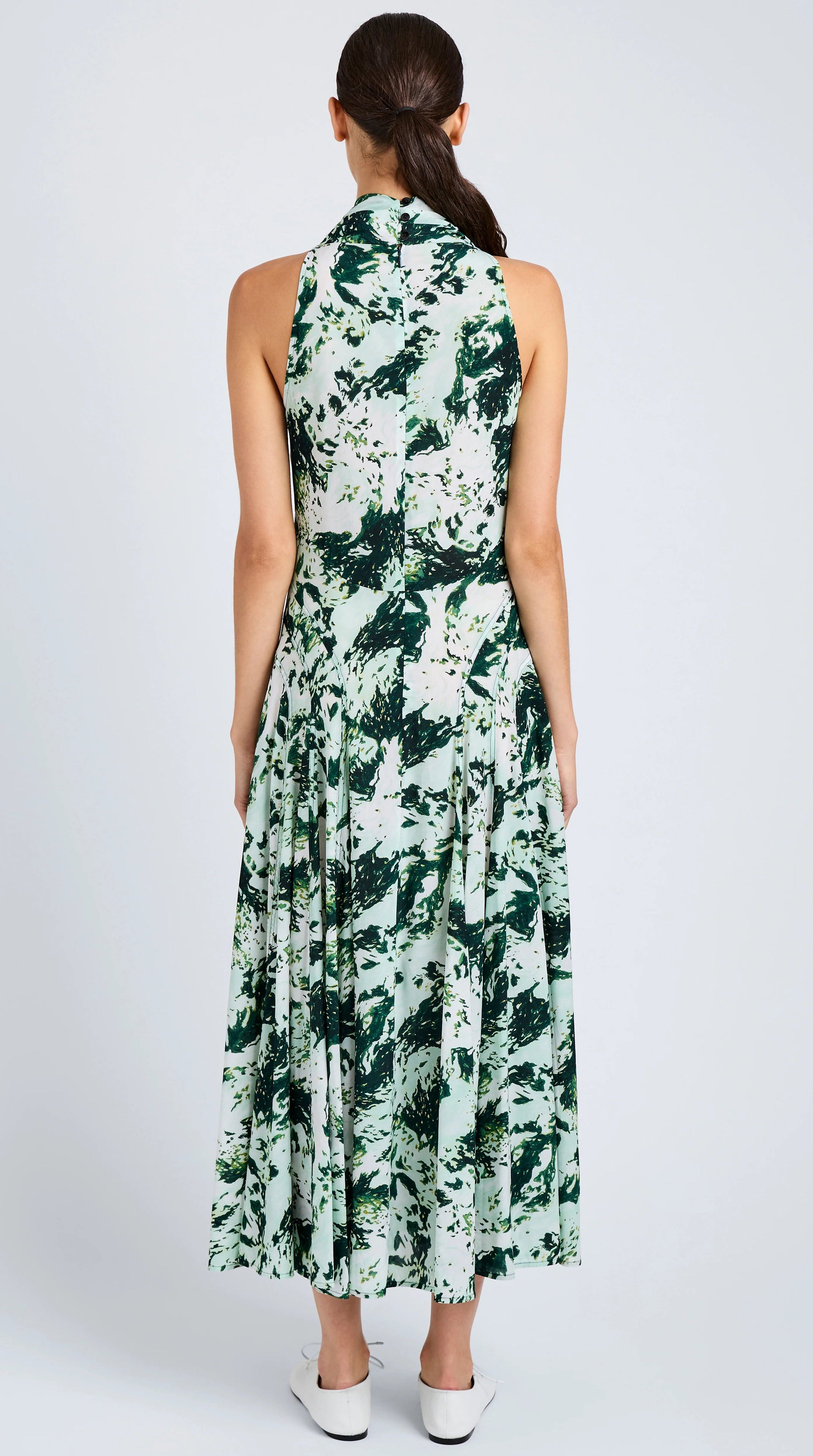 A woman faces away in the Proenza Schouler Adelide Dress—a sleeveless, ankle-length green and white printed silk viscose fit-and-flare dress—paired with white shoes.