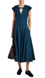 A person wears the Proenza Schouler Manoela Dress—teal, midi, A-line with ruched short sleeves and V-neck cutout—paired with black sneakers and a dark brown tote bag.