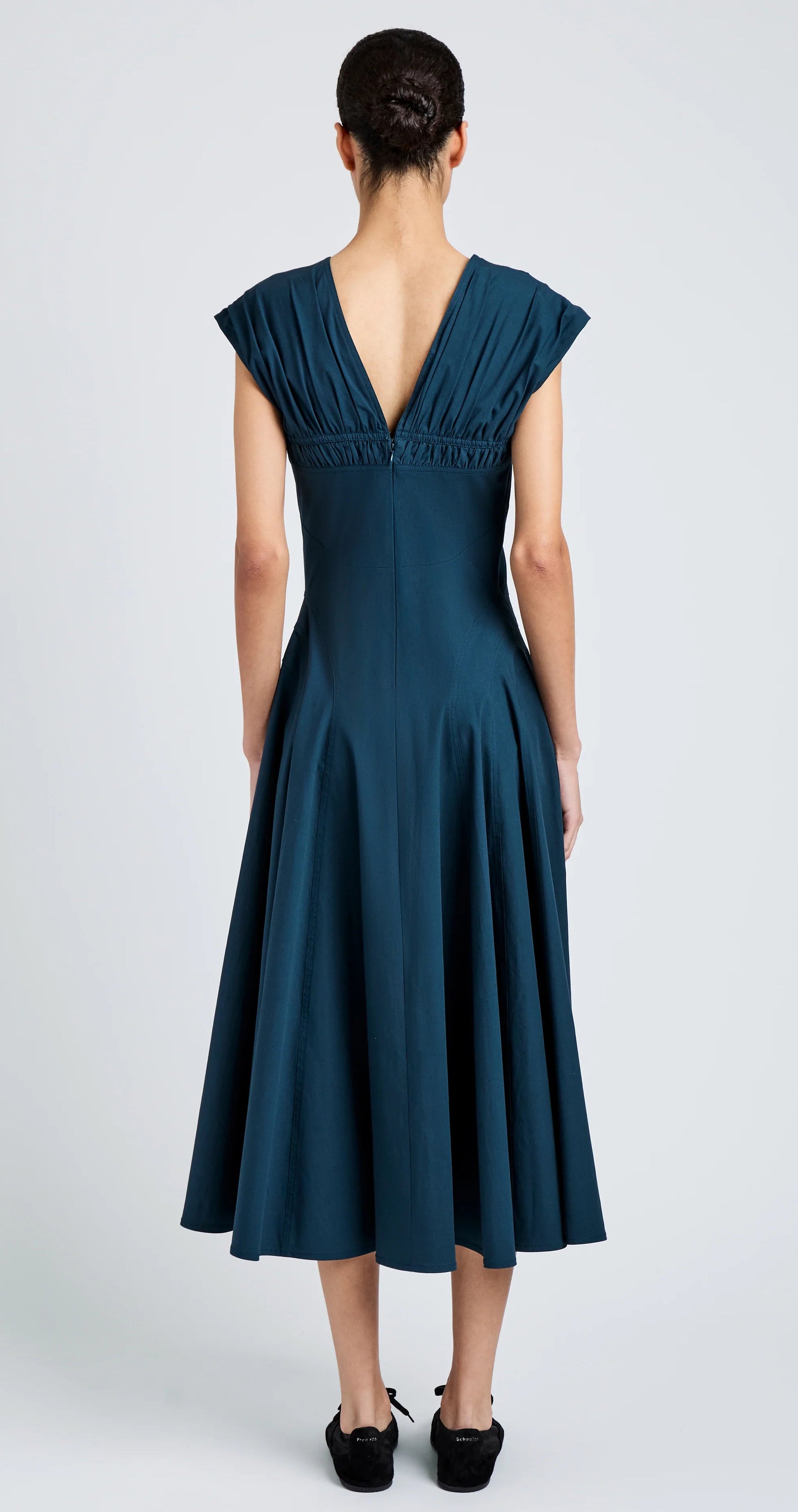 A person faces away, wearing the Manoela Dress by Proenza Schouler—a teal Eco Light Compact Poplin midi with an A-line silhouette, V-back, flared skirt—and black shoes.