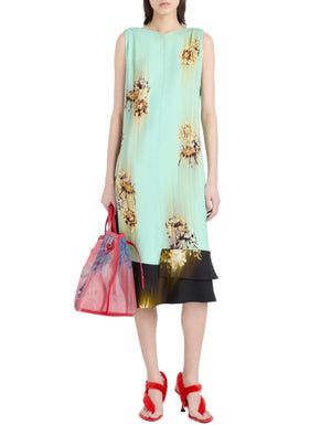 A woman wears the Proenza Schouler Rhetta Dress—sleeveless mint green printed viscose cady with yellow floral ruffle details—paired with a translucent pink bag and red heeled sandals.
