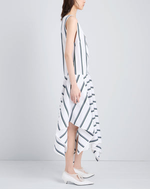 A woman stands sideways in the Proenza Schouler Beatriz Dress, a sleeveless white V-neck dress with black diagonal stripes and an asymmetrical scarf hem, paired with white heeled shoes.