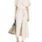 A person wears the Proenza Schouler Isolde Dress, a cream trench-inspired wrap dress with a short cape, paired with a transparent handbag and embellished heeled sandals.