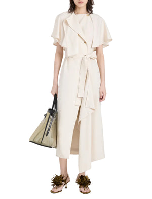 A person wears the Proenza Schouler Isolde Dress, a cream trench-inspired wrap dress with a short cape, paired with a transparent handbag and embellished heeled sandals.