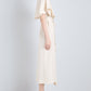 A woman stands in profile wearing the cream Isolde Dress by Proenza Schouler—a trench-inspired, short-sleeve midi style in viscose twill—paired with yellow heeled sandals with floral embellishments.