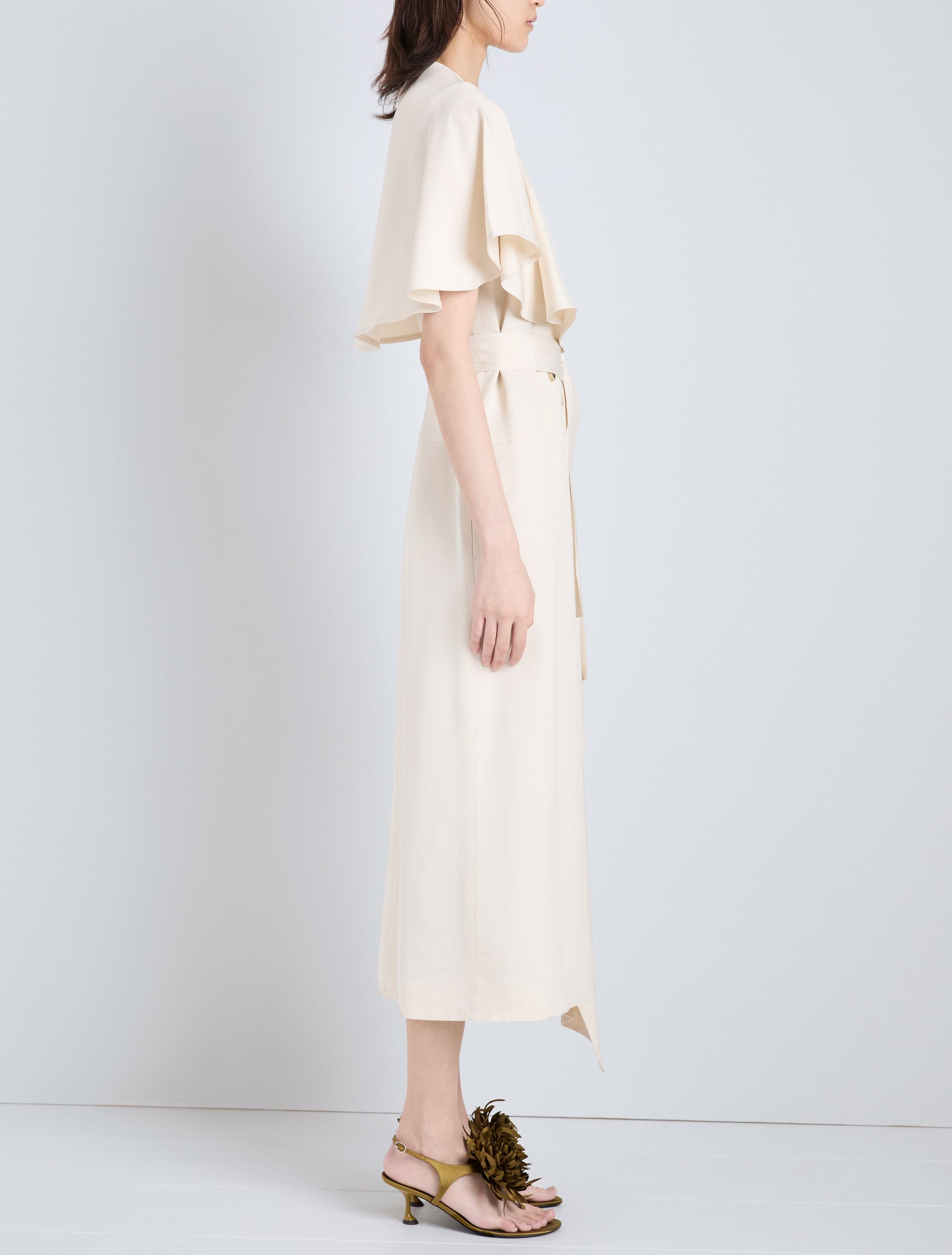 A woman stands in profile wearing the cream Isolde Dress by Proenza Schouler—a trench-inspired, short-sleeve midi style in viscose twill—paired with yellow heeled sandals with floral embellishments.