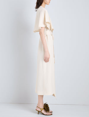 A woman stands in profile wearing the cream Isolde Dress by Proenza Schouler—a trench-inspired, short-sleeve midi style in viscose twill—paired with yellow heeled sandals with floral embellishments.