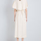 A person stands facing a white wall, wearing the Proenza Schouler Isolde Dress—a cream trench-inspired, belted midi dress with a cape detail in Viscose Twill—paired with open-toe heels.