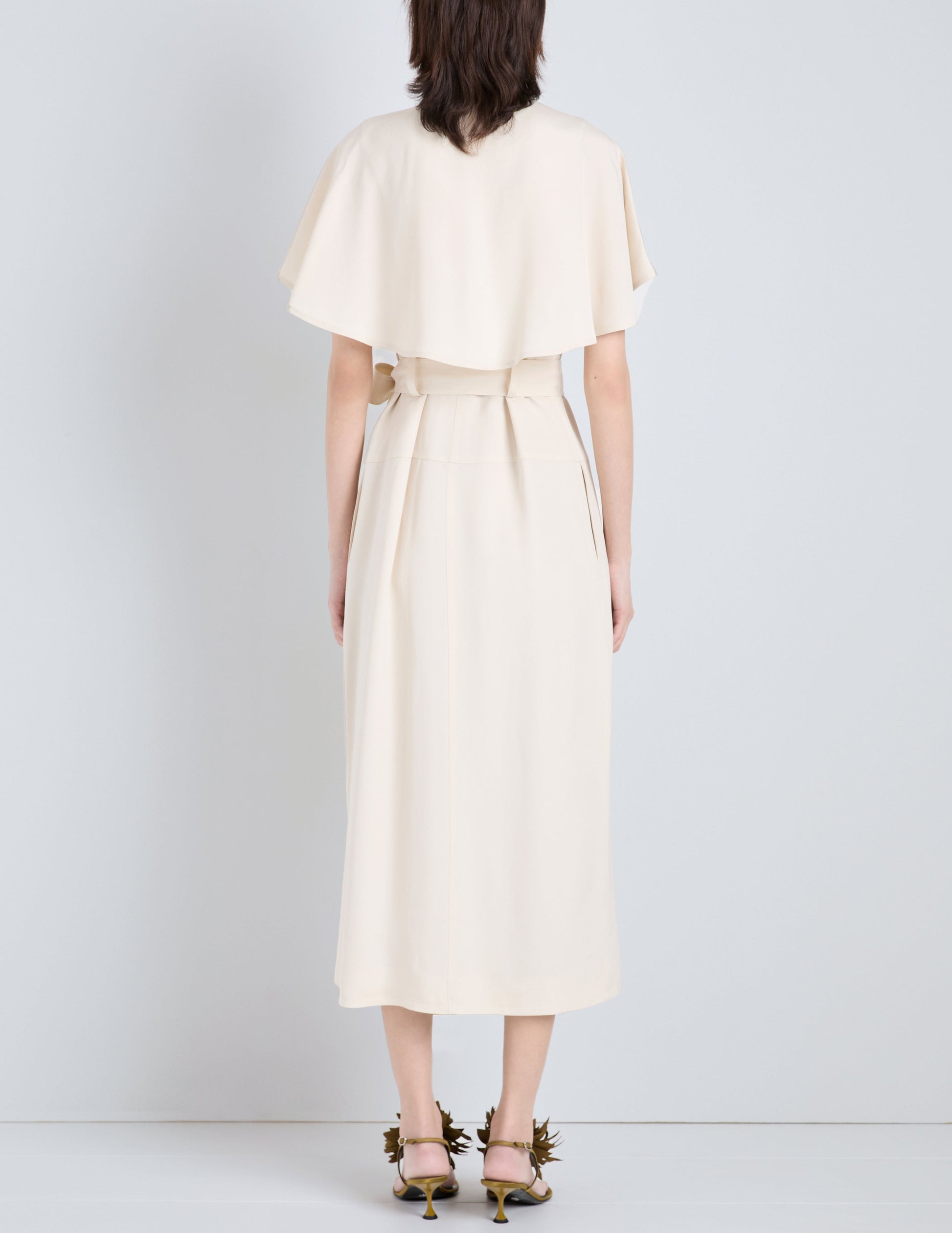 A person stands facing a white wall, wearing the Proenza Schouler Isolde Dress—a cream trench-inspired, belted midi dress with a cape detail in Viscose Twill—paired with open-toe heels.