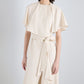 A woman models the Isolde Dress by Proenza Schouler—crafted in cream viscose twill with a cape-style top and wide-leg trousers featuring a tied belt—set against a plain, light background.