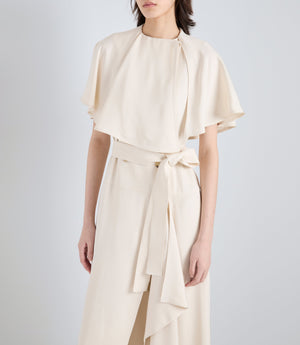 A woman models the Isolde Dress by Proenza Schouler—crafted in cream viscose twill with a cape-style top and wide-leg trousers featuring a tied belt—set against a plain, light background.