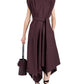 A person wears the Adinna Dress by Proenza Schouler—a sleeveless, collared, dark purple cotton belted dress with an asymmetrical hem—paired with black strappy sandals and a small handbag.