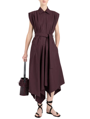 A person wears the Adinna Dress by Proenza Schouler—a sleeveless, collared, dark purple cotton belted dress with an asymmetrical hem—paired with black strappy sandals and a small handbag.