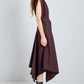 A woman in profile wears the Proenza Schouler Adinna Dress—a sleeveless, dark burgundy dress with an asymmetrical hem and waist belt—paired with black strappy flat sandals against a plain light background.