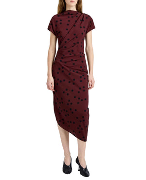 A woman wears the Proenza Schouler Ren Printed Dot Dress, a short-sleeve burgundy midi dress with black polka dots, side draping, and an asymmetrical hem, paired with black heeled shoes.