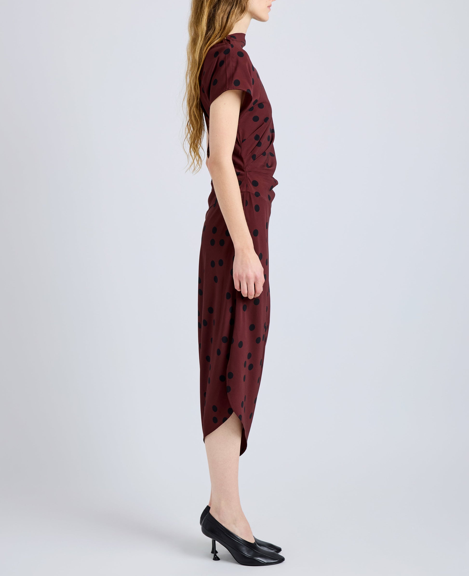 A person stands in profile against a plain light background wearing the Proenza Schouler Ren Printed Dot Dress—maroon with black polka dots, crafted from stretch viscose—paired with black heels.