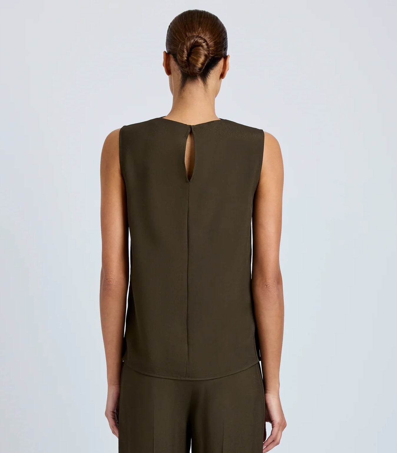 A woman with her hair in a low bun faces away, wearing the Proenza Schouler Defni Top in olive-green Eco Hammered Viscose, featuring a draped high neckline, keyhole back, and matching pants.