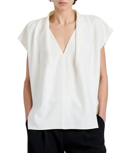 A person is wearing the Proenza Schouler Ranli Top, a loose-fitting, sleeveless white V-neck blouse with a draped collar, paired with black pants and hands in pockets.