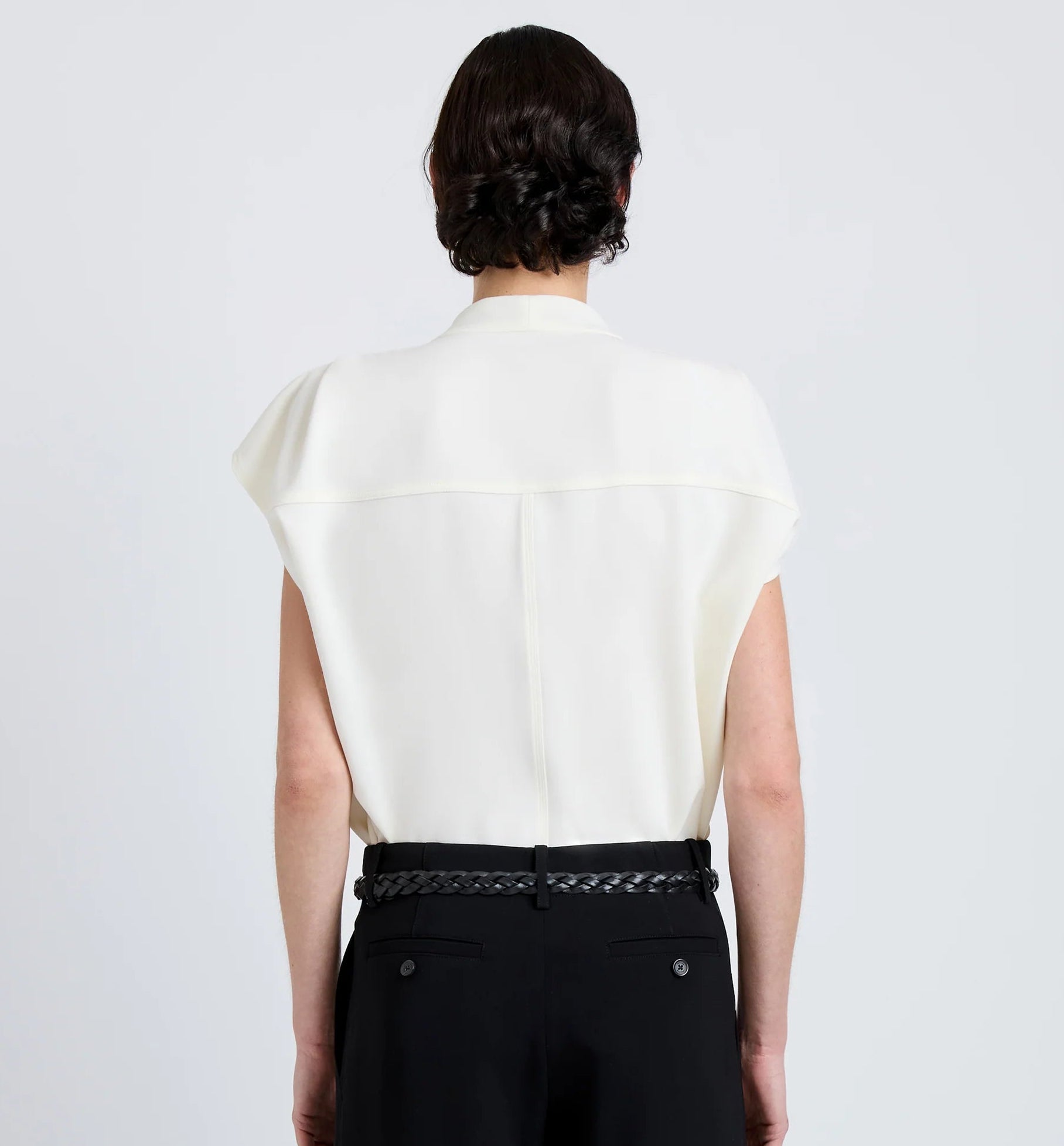 A person with dark hair styled in a low bun is shown from the back, wearing the Proenza Schouler Ranli Top—a white Matte Double Crepe sleeveless blouse with a draped collar—paired with black pants featuring a braided belt, set against a plain light background.