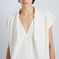 A person is shown wearing the Proenza Schouler Ranli Top, an off-white short-sleeved blouse featuring a draped V-neck collar and crafted from Matte Double Crepe. The image captures the upper body and part of the face against a plain light background.