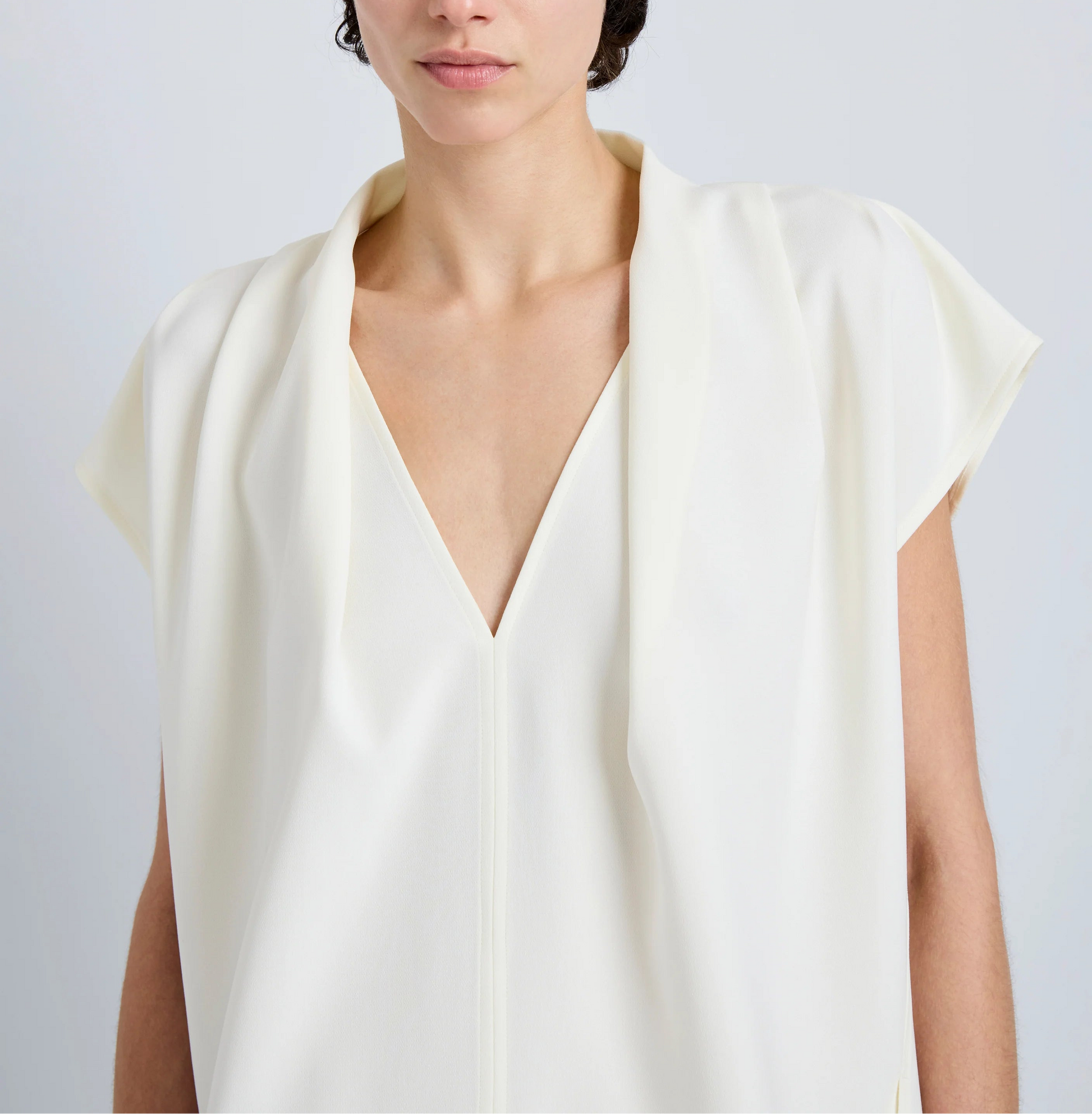 A person is shown wearing the Proenza Schouler Ranli Top, an off-white short-sleeved blouse featuring a draped V-neck collar and crafted from Matte Double Crepe. The image captures the upper body and part of the face against a plain light background.