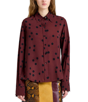 A woman wears the Proenza Schouler Paz Printed Dot Shirt, a maroon stretch viscose top with black polka dots, paired with a snakeskin-patterned skirt featuring yellow and brown panels.