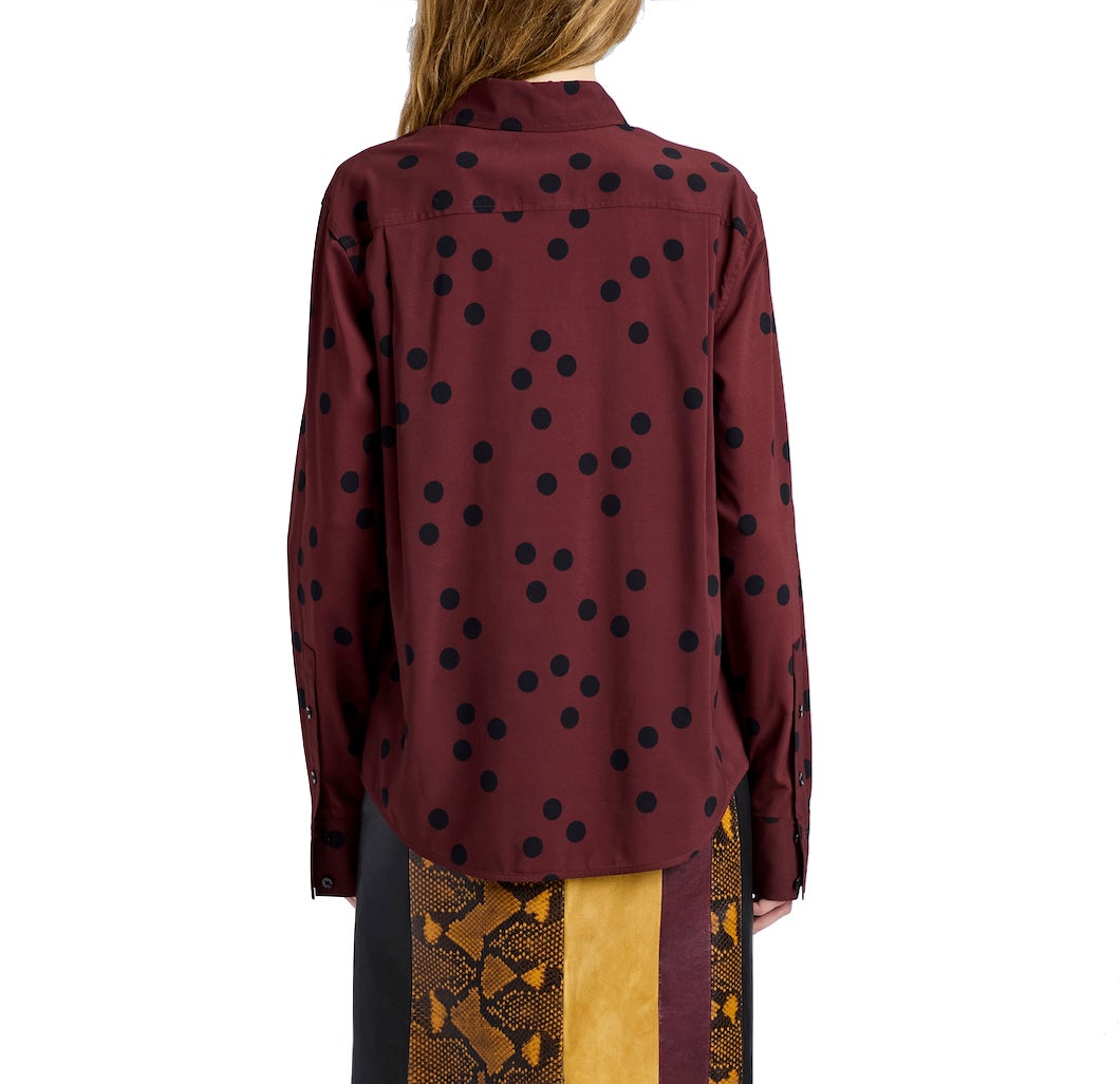 A person is shown from the back wearing the Paz Printed Dot Shirt by Proenza Schouler, a maroon stretch viscose top with black polka dots, paired with a skirt featuring vertical yellow, brown, and black panels.