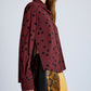 A person wears the Proenza Schouler Paz Printed Dot Shirt—maroon with black polka dots, relaxed fit, side slit, long sleeves, and classic shirting details—paired with an animal print skirt in yellow and brown.