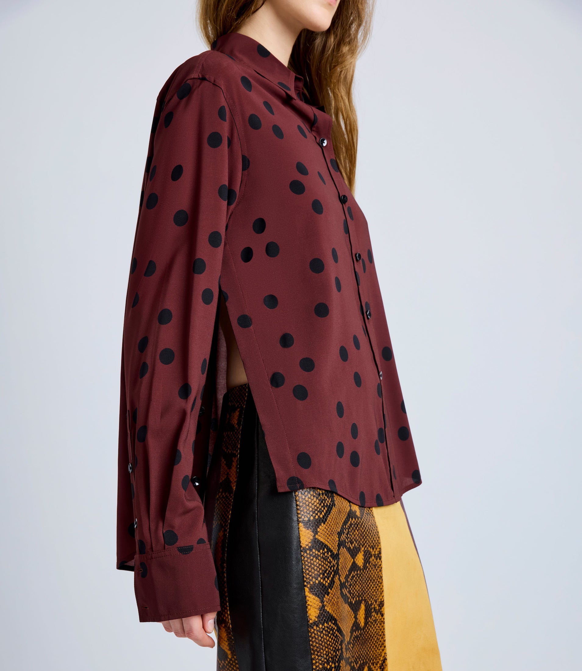 A person wears the Proenza Schouler Paz Printed Dot Shirt—maroon with black polka dots, relaxed fit, side slit, long sleeves, and classic shirting details—paired with an animal print skirt in yellow and brown.