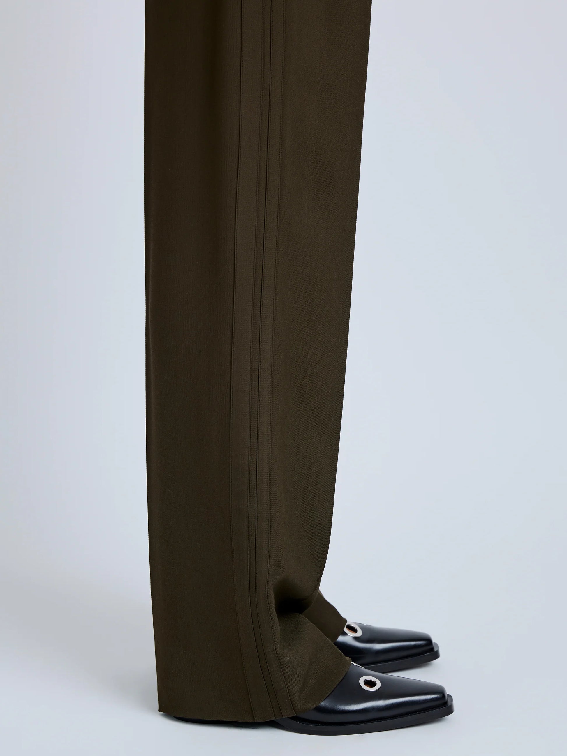 Someone wears the Proenza Schouler Lien Pant in dark olive, a high-waisted wide-leg style in eco hammered viscose that covers most of black square-toe shoes with metallic ring detail, set against a light neutral background.