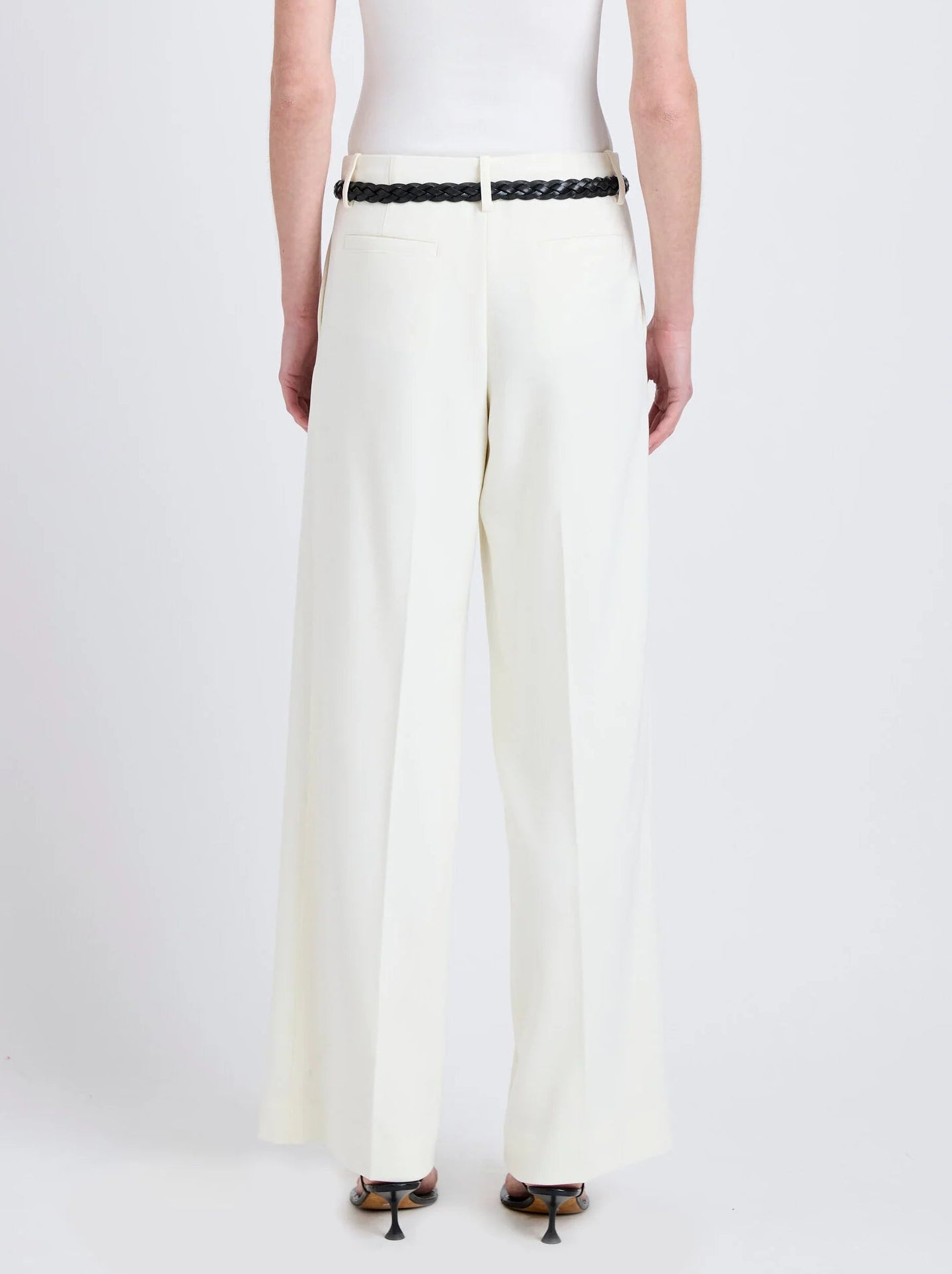 A person wearing the Proenza Schouler Halima Pant in white high-waisted matte double crepe, paired with a white sleeveless top, black braided belt, and heeled sandals, stands facing away against a plain background.