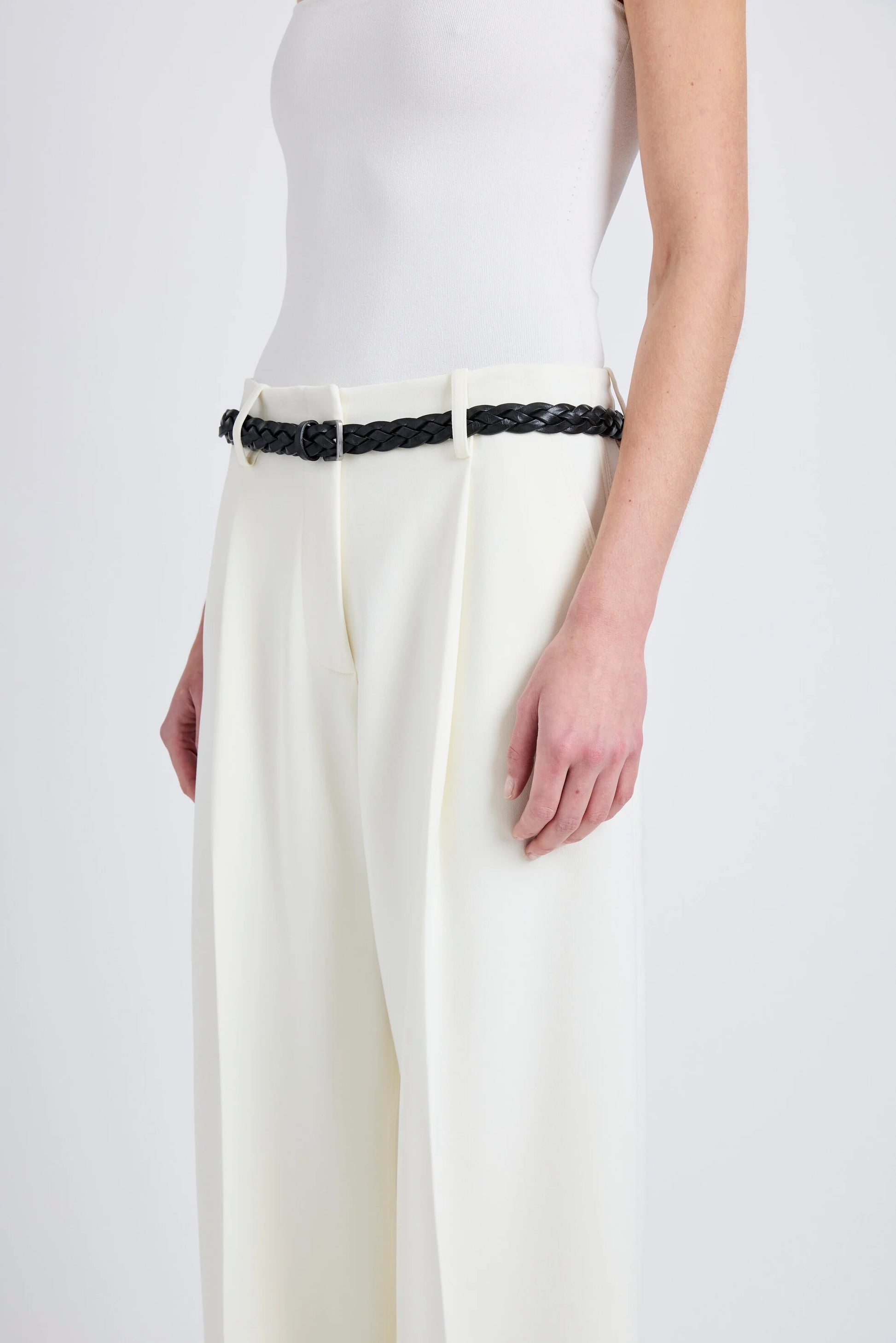 A person shown from shoulders to mid-thigh against a plain background wears a sleeveless white top, the Proenza Schouler Halima Pant in matte double crepe with a high waist, and a black braided belt.