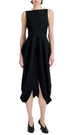 A person wearing the Sukki Dress by Proenza Schouler, a sleeveless black draped viscose dress with a fitted, backless bodice, peplum waist, and asymmetrical skirt, paired with black heels.