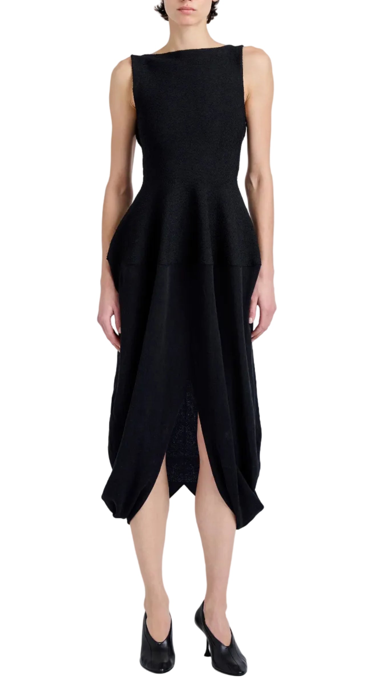 A person wearing the Sukki Dress by Proenza Schouler, a sleeveless black draped viscose dress with a fitted, backless bodice, peplum waist, and asymmetrical skirt, paired with black heels.