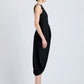 A woman in profile wears the Sukki Dress by Proenza Schouler—a sleeveless black viscose design with a fitted, backless bodice and draped skirt—paired with black heels against a plain light background.