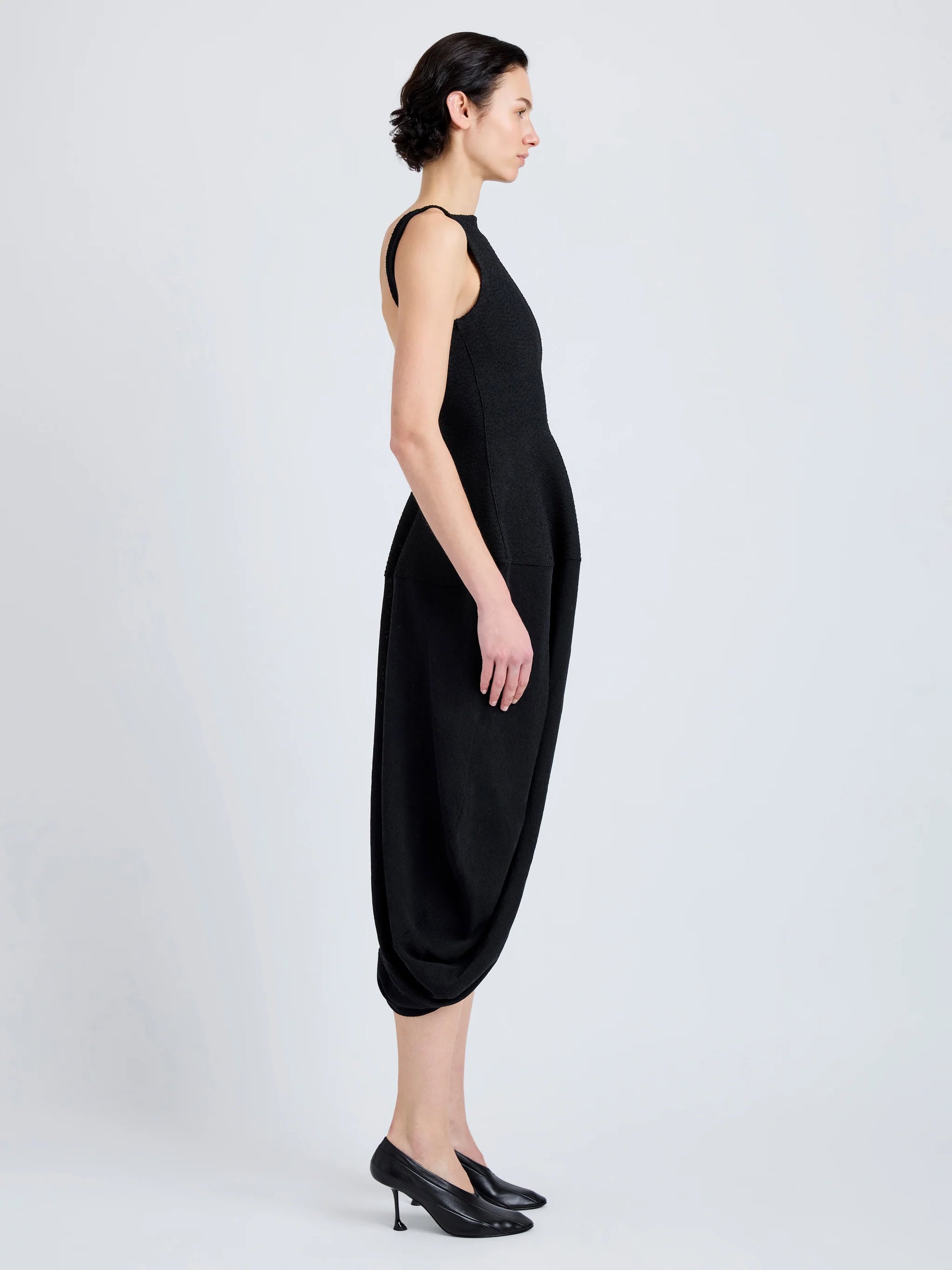 A woman in profile wears the Sukki Dress by Proenza Schouler—a sleeveless black viscose design with a fitted, backless bodice and draped skirt—paired with black heels against a plain light background.