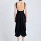 A person faces away, wearing the Sukki Dress by Proenza Schouler—a sleeveless black draped viscose dress with a low-cut backless bodice and loose calf-length skirt—paired with black heels against a plain background.
