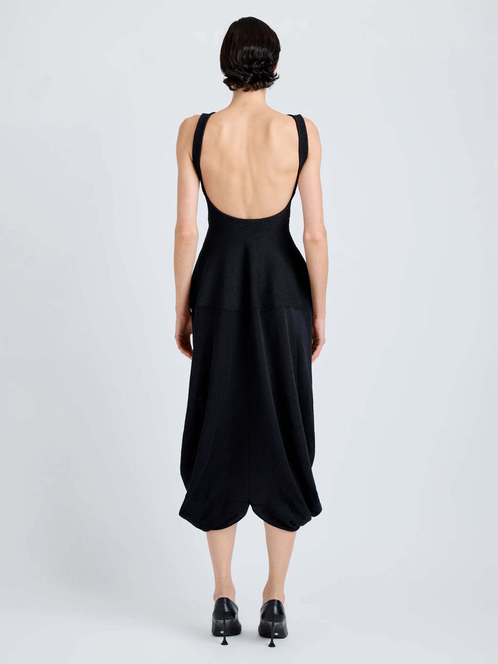 A person faces away, wearing the Sukki Dress by Proenza Schouler—a sleeveless black draped viscose dress with a low-cut backless bodice and loose calf-length skirt—paired with black heels against a plain background.