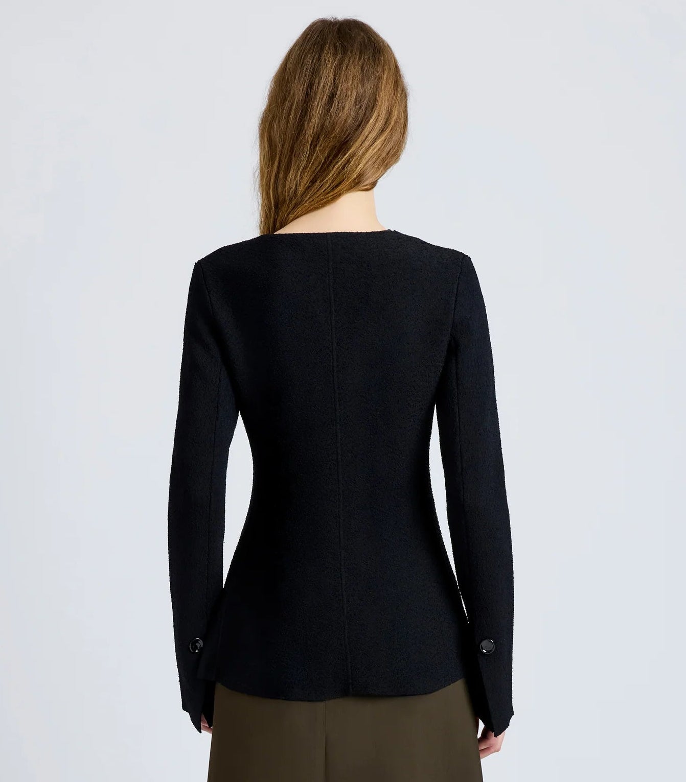 A woman with long brown hair is seen from behind wearing the Sukki Jacket by Proenza Schouler in black, paired with an olive green skirt against a plain light background.