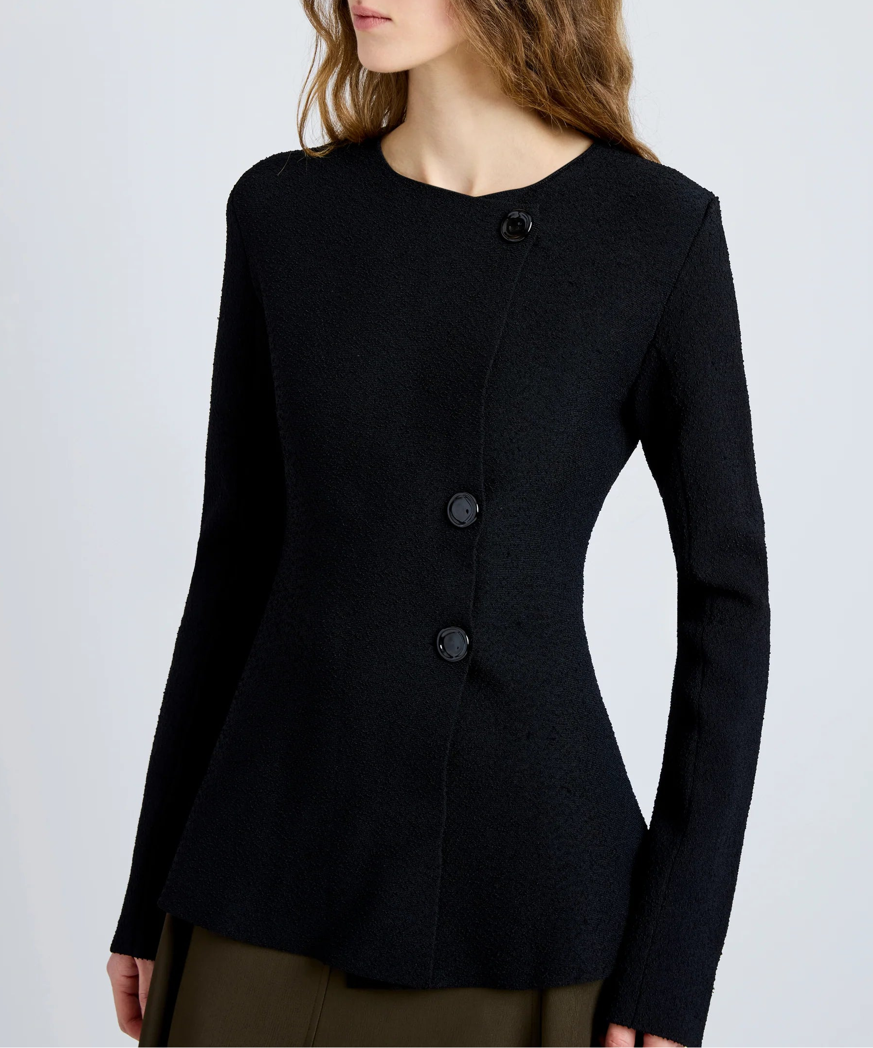 A woman wears the Sukki Jacket by Proenza Schouler, a tailored black jacket with an asymmetric front and three large black buttons, paired with olive green pants.