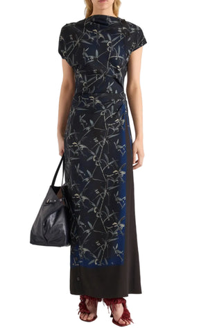 A woman stands against a white background wearing the Proenza Schouler Anais Dress in navy and black Brazilian night orchids floral print, crafted from printed stretch cady. She carries a black handbag and completes her look with red fringed heels.