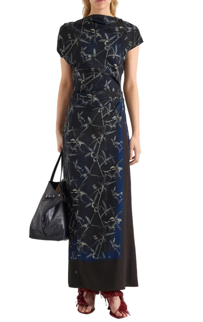 A woman stands against a white background wearing the Proenza Schouler Anais Dress in navy and black Brazilian night orchids floral print, crafted from printed stretch cady. She carries a black handbag and completes her look with red fringed heels.