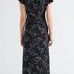 A person stands with their back to the camera, dressed in the Proenza Schouler Anais Dress—an ankle-length black dress featuring a gray Brazilian night orchids pattern on Printed Stretch Cady with short sleeves—paired with red heeled shoes.