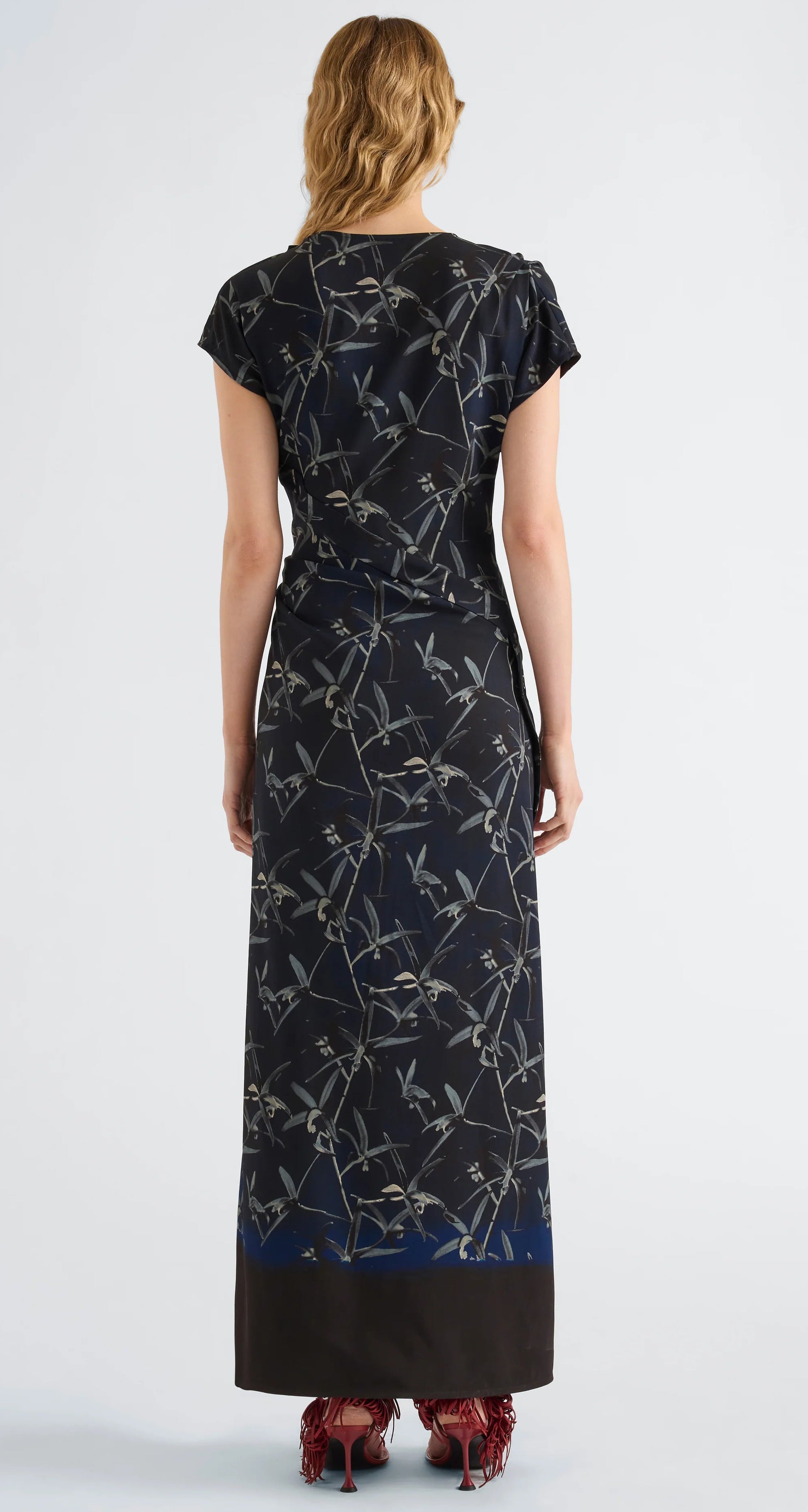 A person stands with their back to the camera, dressed in the Proenza Schouler Anais Dress—an ankle-length black dress featuring a gray Brazilian night orchids pattern on Printed Stretch Cady with short sleeves—paired with red heeled shoes.