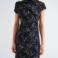 Wearing the Anais Dress by Proenza Schouler—a short-sleeved, navy blue design fashioned from printed stretch cady and embellished with delicate gray Brazilian night orchid motifs—a woman stands before a plain background.