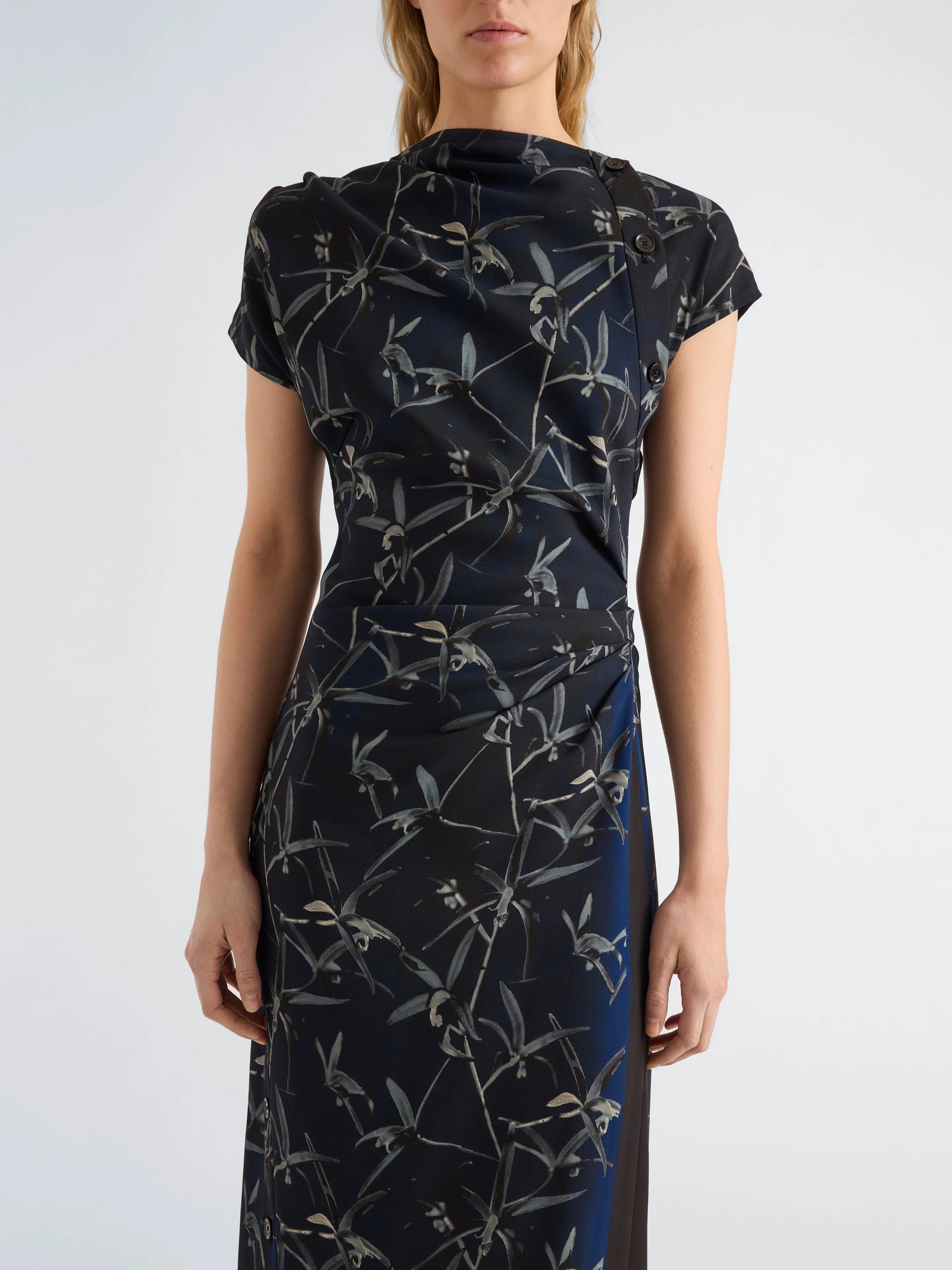 Wearing the Anais Dress by Proenza Schouler—a short-sleeved, navy blue design fashioned from printed stretch cady and embellished with delicate gray Brazilian night orchid motifs—a woman stands before a plain background.