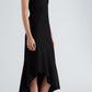 A woman stands against a plain background, modeling the Proenza Schouler Elodine Tropical Wool Dress—a sleeveless, high-neck, asymmetrical black midi crafted from lightweight wool—highlighting its sleek transitional silhouette paired with black pointed-toe shoes.
